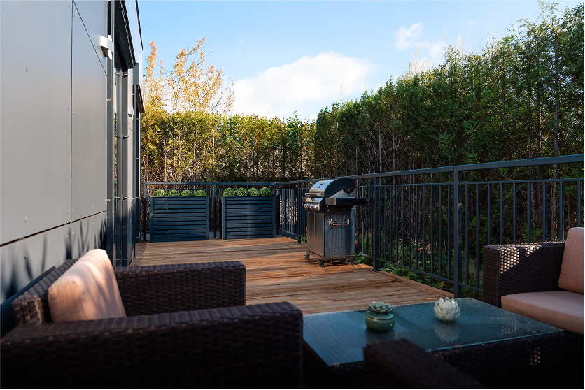 Terrasse beim 2B Mikrohaus mit Lounge-Möbeln, Holzboden und Grill, umgeben von Hecken.